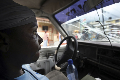 A woman passenger rides in a shared taxi. The taxi's windshield is damaged and the woman has an anxious expression on her face.
